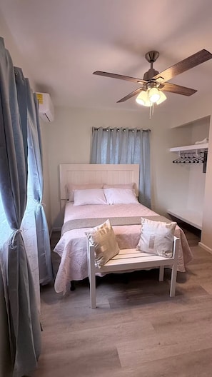 Ein Schlafzimmer mit einem Bett, einem Deckenventilator, einem Fenster mit Vorhängen und einem an der Wand befestigten Regal.