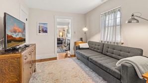 A modern living room with a grey sofa, a flat-screen TV, a wooden cabinet, a patterned rug, and a large window with sheer curtains. - Route 66/Grand Canyon Retreat, Central Williams, Family-friendly, Sleeps 8 (Williams)