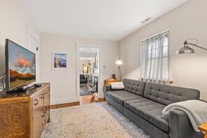 A modern living room with a grey sofa, a flat-screen TV, a wooden cabinet, a patterned rug, and a large window with sheer curtains. - Route 66/Grand Canyon Retreat, Central Williams, Family-friendly, Sleeps 8 (Williams)
