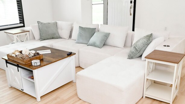 Een witte zithoek met een houten salontafel en bijzettafeltje, een witte kast met een houten blad, en een raam met jaloezieën.