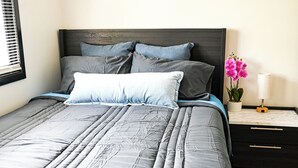 Een bed met grijze beddengoed, een donker hoofdeinde en een nachtkastje met een vaas roze bloemen.