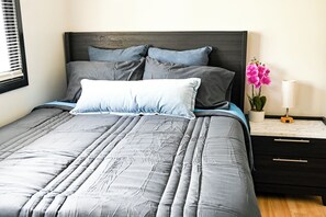 Ein Bett mit grauer Bettwäsche, ein dunkles Kopfteil und ein Nachttisch mit einer Vase voller pinkfarbener Blumen.