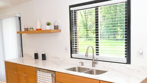 Een keuken met een dubbele spoelbak, houten kastjes, een marmeren aanrechtblad, een raam met jaloezieën en een plank met decoratieve objecten.