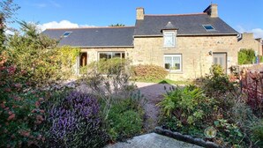 Une maison en pierre avec une allée pavée, un petit jardin agrémenté de diverses plantes et un ciel d’un bleu éclatant.