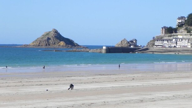 Une plage avec un promontoire rocheux, une mer calme et des bâtiments situés sur une falaise.