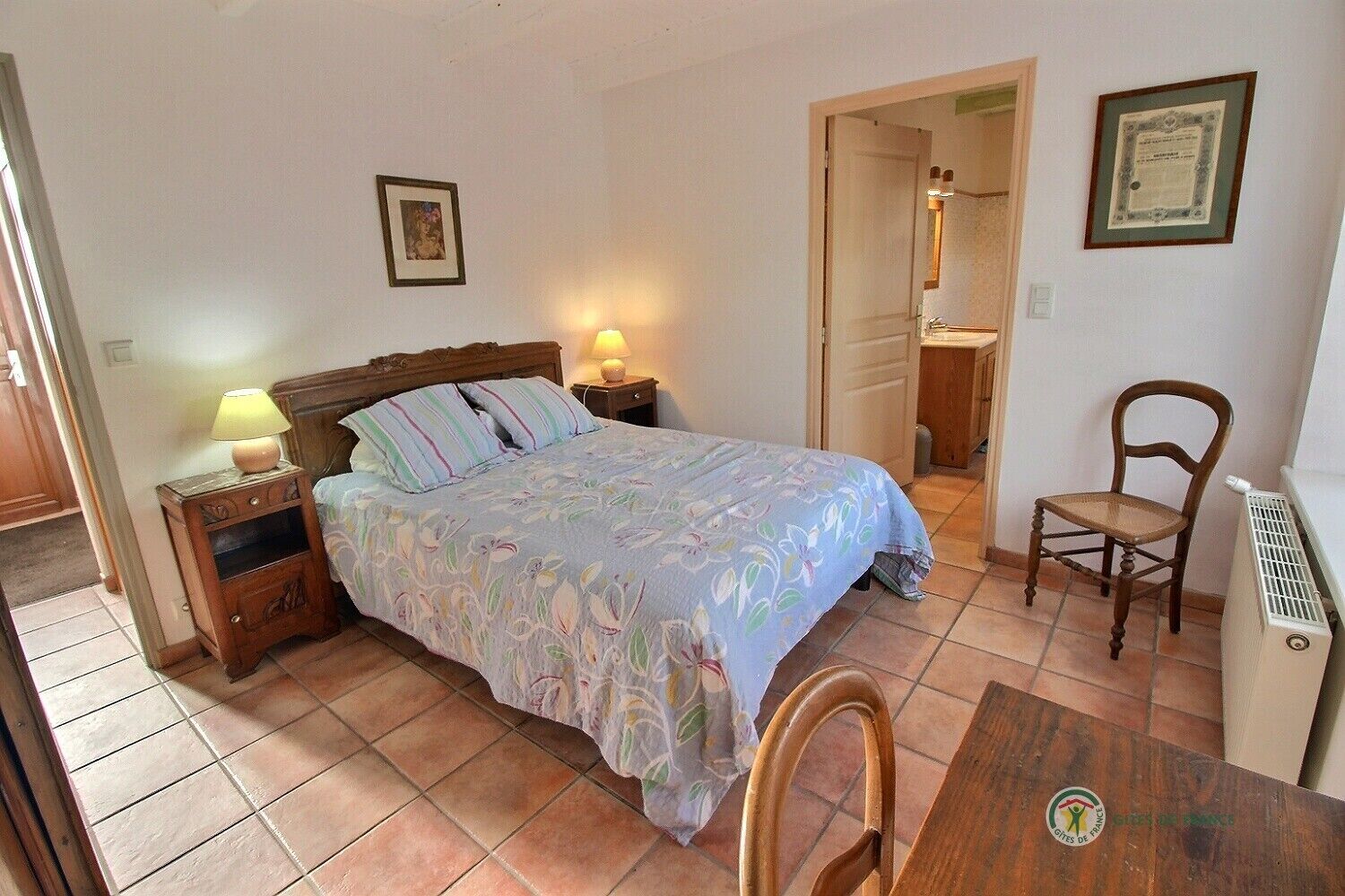 Un dormitorio con cama, lámparas de noche, una silla, un radiador y un certificado enmarcado en la pared.