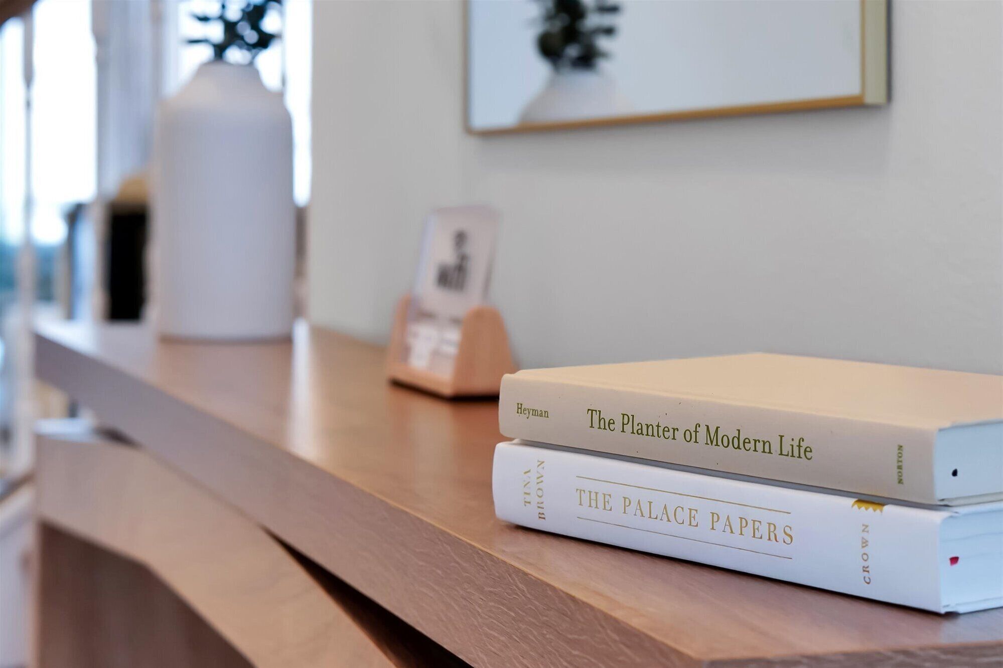 A stack of books on a wooden surface with a framed picture in the background.