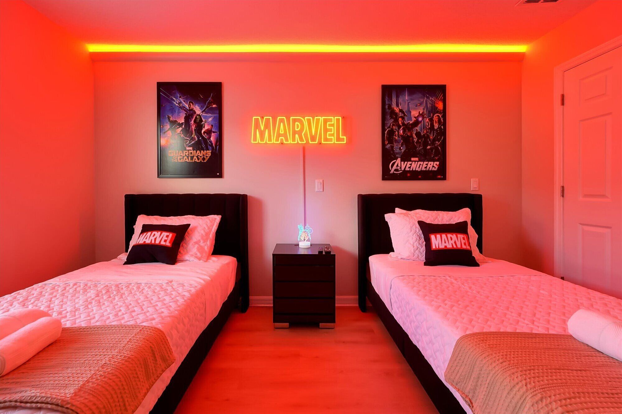 A bedroom with two beds, each with a pillow featuring the Marvel logo, a neon sign with the word 'MARVEL', and posters of 'Guardians of the Galaxy' and 'Avengers' on the wall.