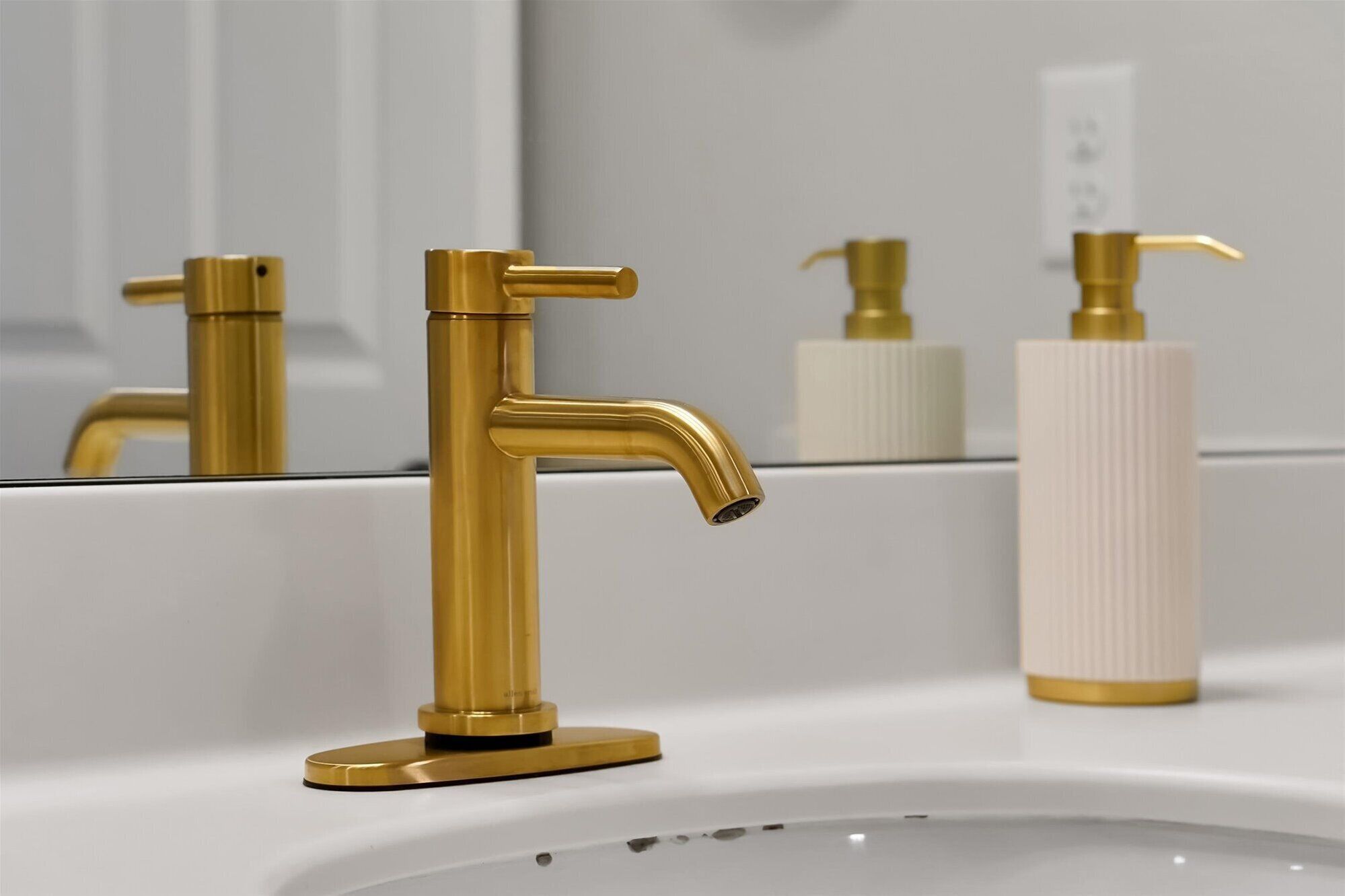 Un baño con grifo dorado, dispensador de jabón y un espejo que refleja el lavamanos.