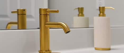 Un baño con grifo dorado, dispensador de jabón y un espejo que refleja el lavamanos.