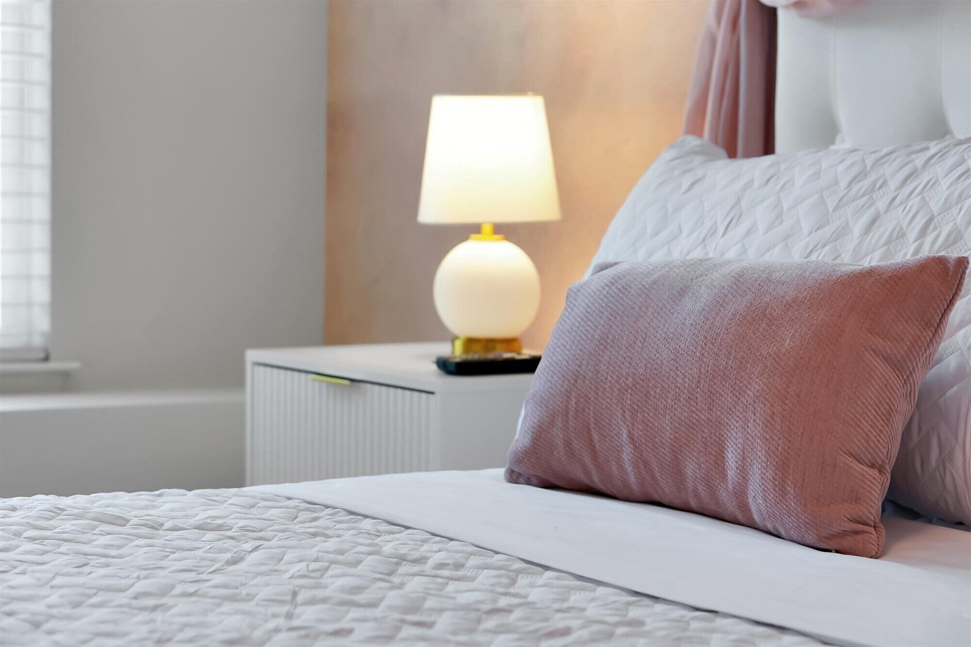 Een net opgemaakt bed met een witte deken, een roze kussen, een bedlampje en een witte nachtkastje.