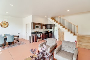 A modern kitchen with dark cabinets, a stainless steel refrigerator, and a microwave. There is a dining area with a table and chairs, and a staircase leading to an upper level.