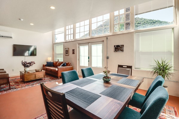 A spacious living area with a dining table, comfortable seating, a flat-screen TV, and large windows offering a view of the outdoors.