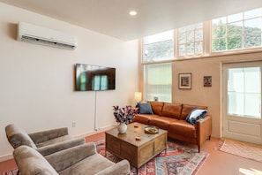 A living room with a brown leather sofa, a grey armchair, a wooden coffee table, a TV mounted on the wall, and a large window with blinds.