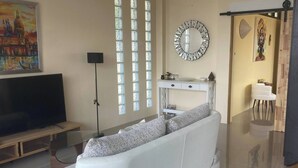 Een moderne woonkamer met een witte bank, een flatscreen televisie, een groot raam met verticale jaloezieën en een decoratieve spiegel.