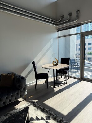 Une pièce moderne avec un canapé, une table ronde et des chaises, de grandes fenêtres donnant sur des immeubles et des conduits apparents au plafond.