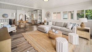 En rummelig opholdsstue med sofa, skammel, sofabord og et tæppe. Der er et tekøkken med et spisebord og stole. Væggene er hvide og gulvene er i træ.