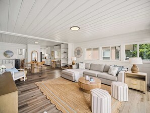 A spacious living area with a sofa, ottoman, coffee table, and a rug. There is a kitchen area with a dining table and chairs. The room has white walls and wooden flooring. - Little Hamptons on The Lake (Newport)