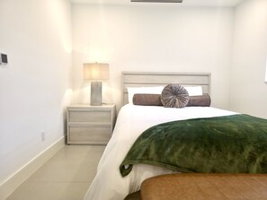 A bedroom with a bed, a nightstand with a lamp, a drawer, and a bench. - Coral Springs Oasis Retreat (Coral Springs)