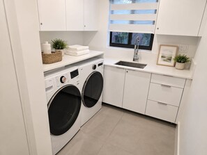 A modern laundry area with a front-loading washer and dryer, a sink, and a window with blinds. - Coral Springs Oasis Retreat (Coral Springs)