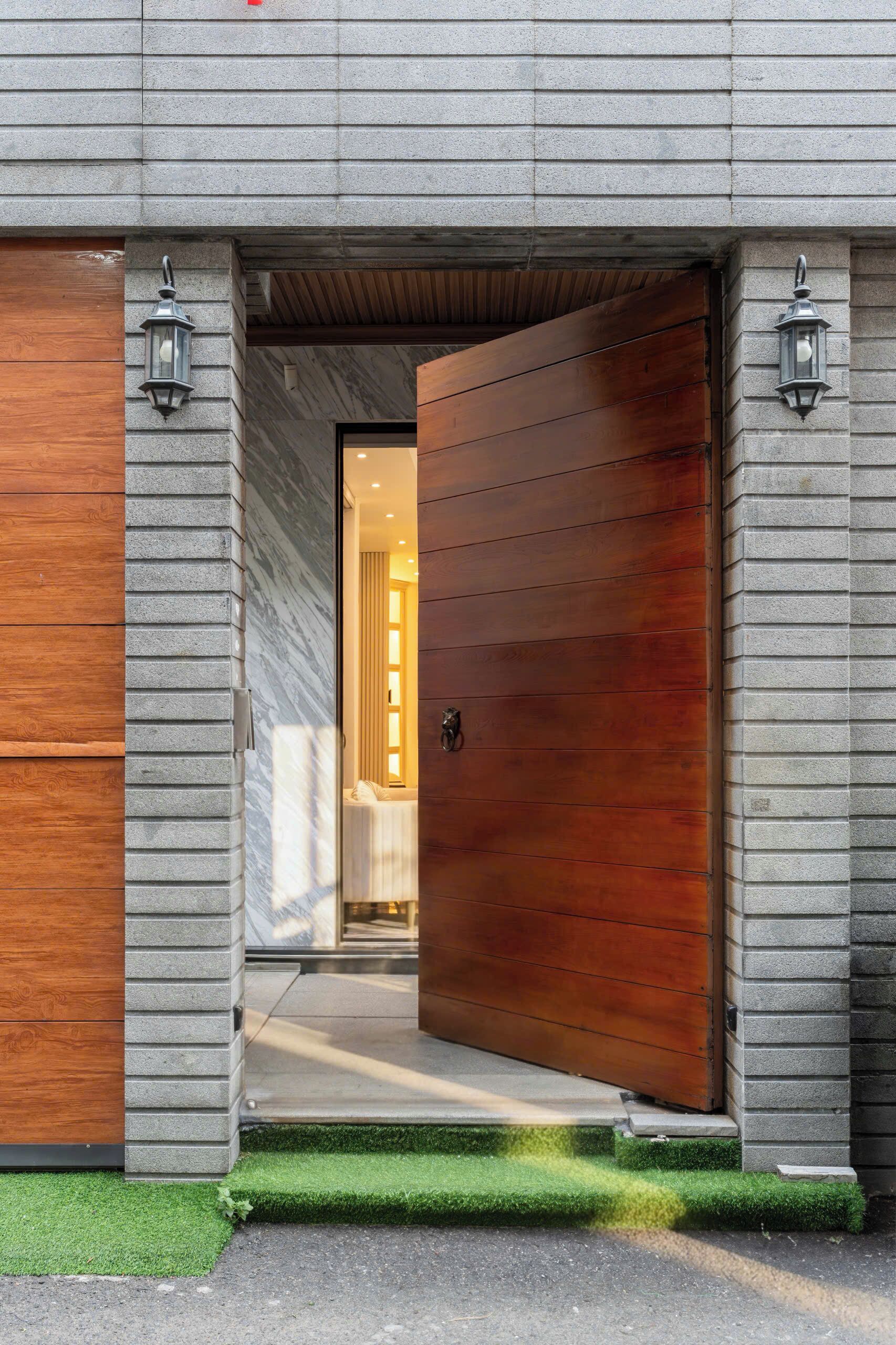 A modern building entrance with a large wooden door, two wall-mounted lanterns, and a pathway leading to a well-lit interior.