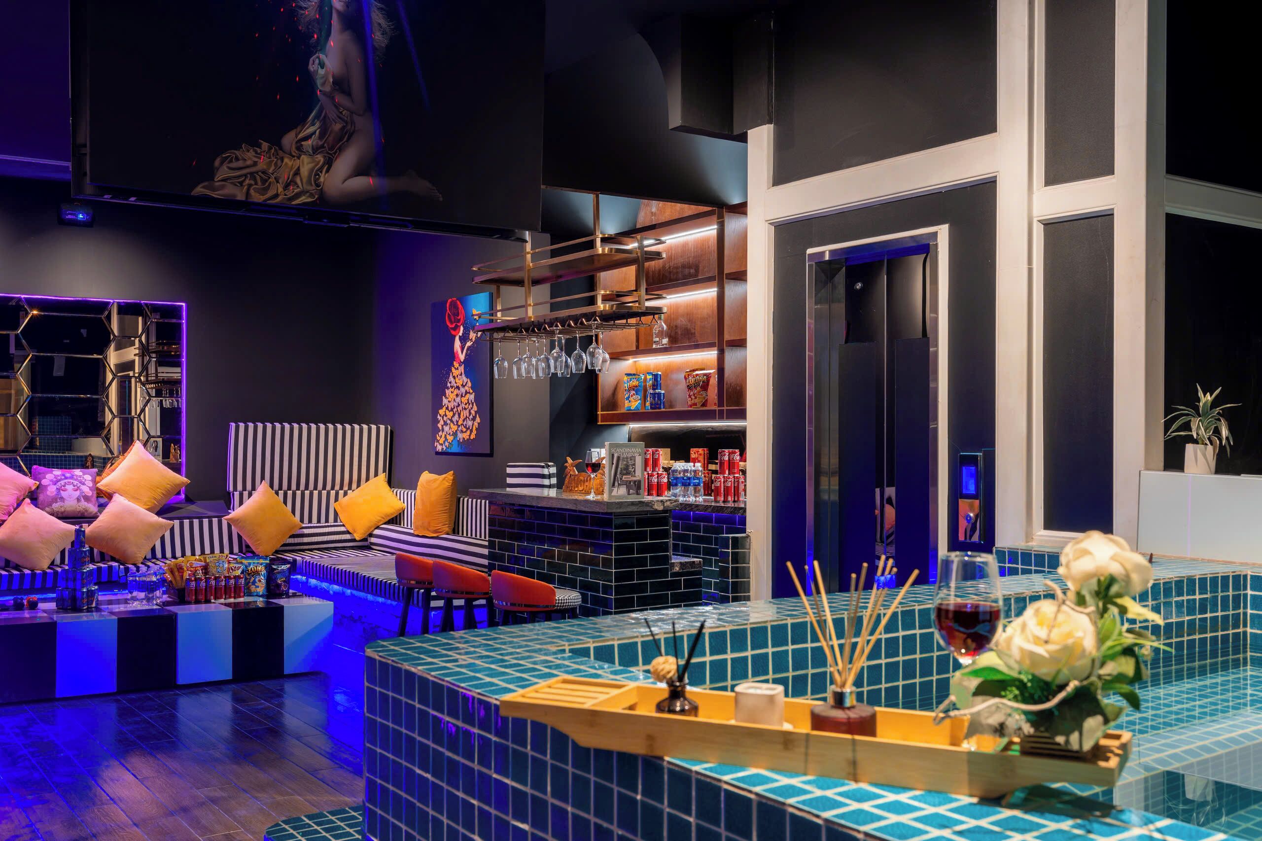 A modern bar area with a blue-tiled bar counter, seating with colorful cushions, and a large screen above.