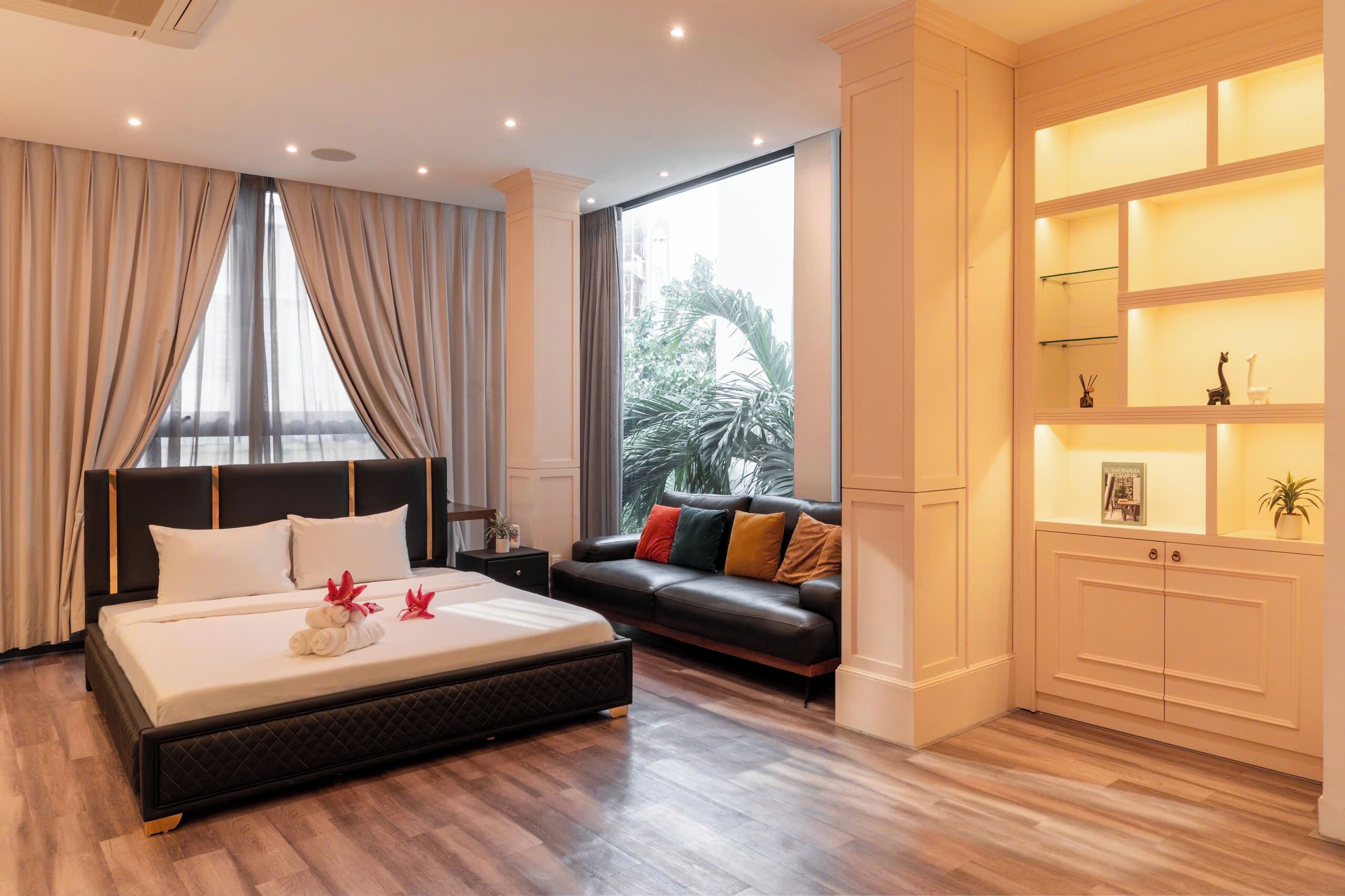 A modern hotel room with a large bed, a sofa, a nightstand, and a built-in shelf with decorative items.