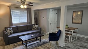 A living room with a sofa, coffee table, and a chair. There's a ceiling fan, a door, and a framed map on the wall.