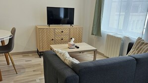 A modern living room with a sofa, a wooden cabinet with a TV, a table with a remote, and a window with curtains. - Studio Cosy Centre Valenciennes Station and streetcar on foot (Valenciennes)