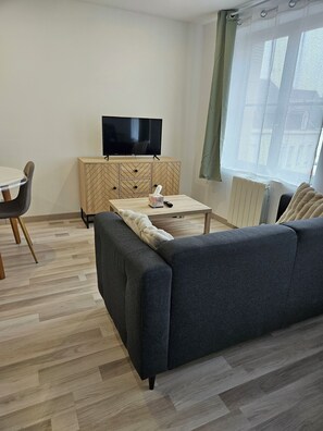 A modern living room with a sofa, a wooden cabinet with a TV, a table with a remote, and a window with curtains. - Studio Cosy Centre Valenciennes Station and streetcar on foot (Valenciennes)