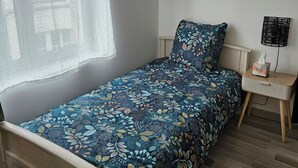A single bed with a floral bedspread, a bedside table with a tissue box, a power outlet, and a window with sheer curtains. - Studio Cosy Centre Valenciennes Station and streetcar on foot (Valenciennes)