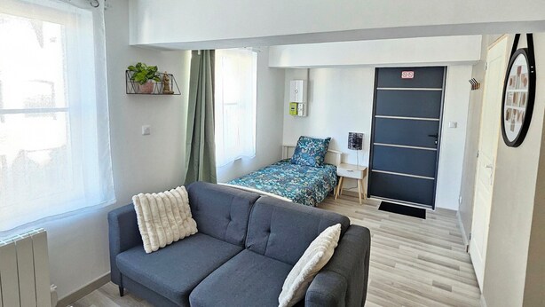 A modern apartment with a grey sofa, a bed with floral bedding, a radiator, a white desk, and a door with a sign. - Studio Cosy Centre Valenciennes Station and streetcar on foot (Valenciennes)