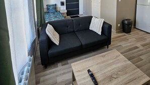 A modern living room with a black sofa, a wooden coffee table, a remote control, a kitchen area with a refrigerator, and a bed with a blue and white patterned bedspread. - Studio Cosy Centre Valenciennes Station and streetcar on foot (Valenciennes)