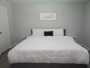 A bedroom with a bed, a bedside table, a nightstand, a framed quote on the wall, and a door. - Gateway City Getaway (Forest Park)