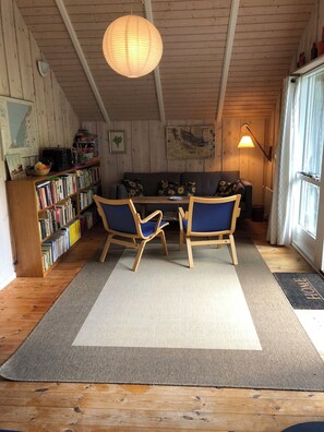 Ein gemütliches Zimmer mit Holzbalkendecke, einer Bücherwand, einem Sofa mit Sonnenblumen und einem Essbereich mit Stühlen.