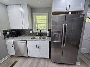 A compact kitchen with white cabinets, a stainless steel refrigerator, a built-in water dispenser, and a window with blinds. - Silver Group Rentals Presents The Silver Spot (Atlanta)