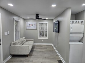 A hallway with a gray sofa, a ceiling fan, a wall-mounted TV, and a stacked washer and dryer. - Silver Group Rentals Presents The Silver Spot (Atlanta)
