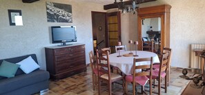 Un coin repas avec une table dressée pour quatre personnes, un téléviseur posé sur une commode, un canapé avec des coussins et un miroir qui reflète la pièce.