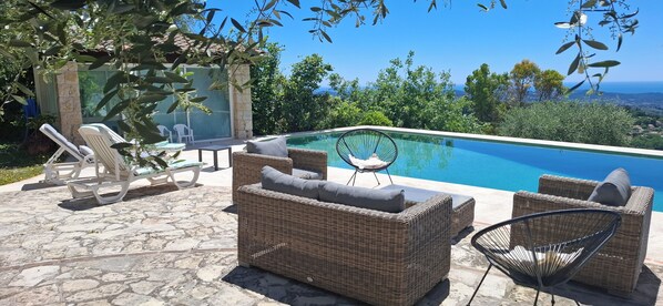 Un espace amĂ©nagĂ© au bord de la piscine, avec des meubles en osier, une structure fermĂ©e par des parois de verre et une vue sur lâocĂ©an.