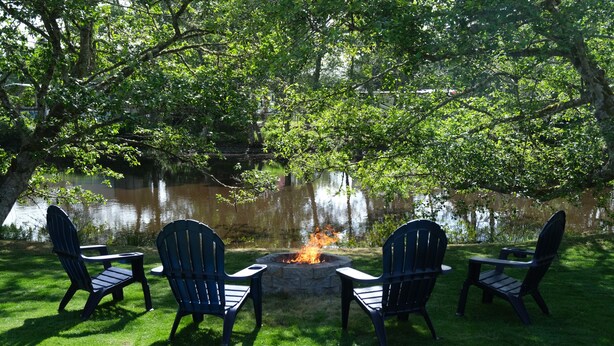 Een vuurplaats met vier Adirondack-stoelen eromheen, gelegen in een grasrijk gebied met bomen en een waterlichaam op de achtergrond.