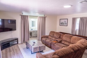 Ein Wohnzimmer mit einer Eckcouch, einem Flachbildfernseher, einem Couchtisch und einer Küche im Hintergrund.