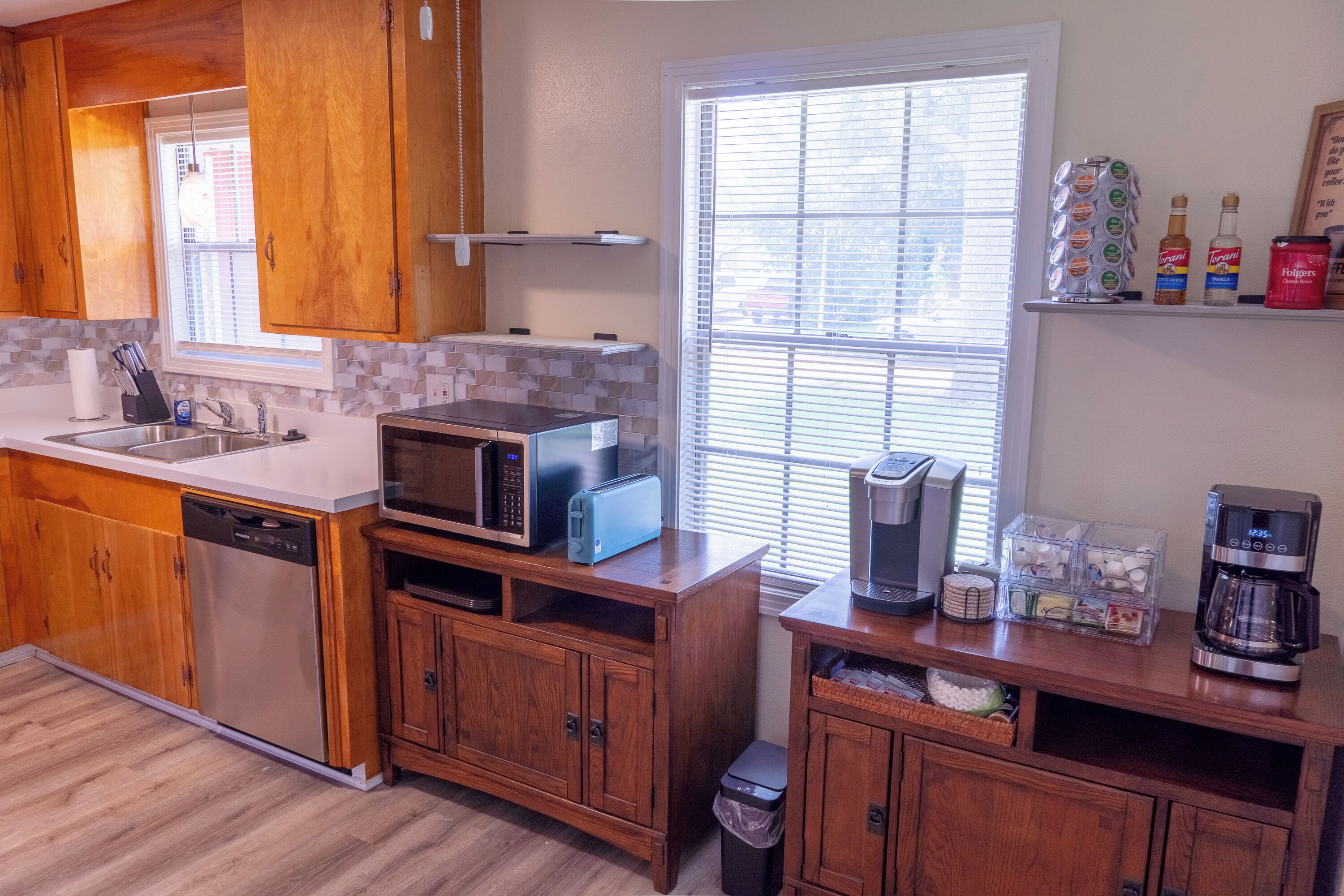 A kitchen with wooden cabinets, a dishwasher, a microwave, a toaster, a coffee maker, and a window with blinds.