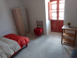 Une chambre Ă coucher avec un lit, une chaise, une armoire, une petite table et une fenĂȘtre avec des rideaux.
