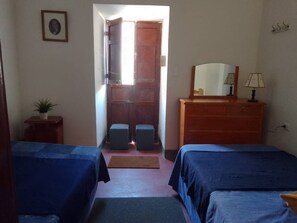 A room with two beds, a dresser, a mirror, a lamp, a small table with a plant, and a door leading outside.