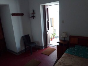 Una habitació amb un llit, una cadira, un escriptori, una làmpada i una porta que dona a zona exterior amb plantes en testos.