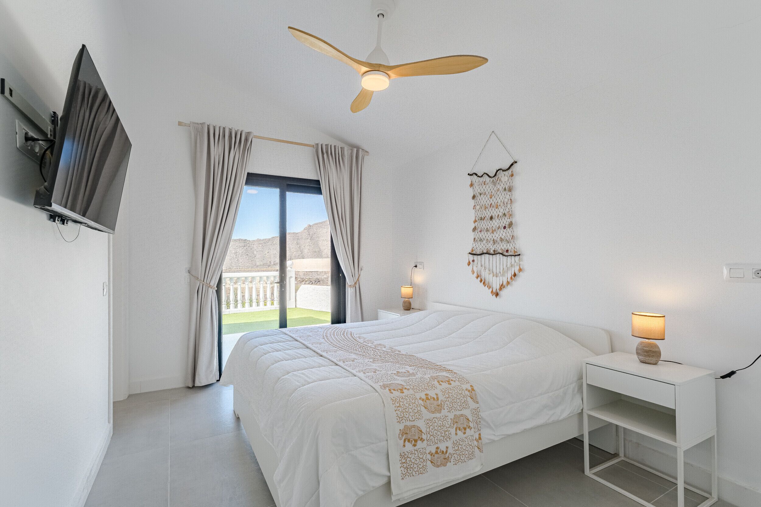 A bedroom with a bed, a ceiling fan, a TV mounted on the wall, a bedside table with a lamp, and a view of the outdoors.