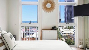 A bedroom with a bed, a radiator, a wall-mounted TV, and a view of buildings through large windows. - 1 bd apartment - Heated Pool, Spa, Walk to the Beach | Broadbeach (Broadbeach)