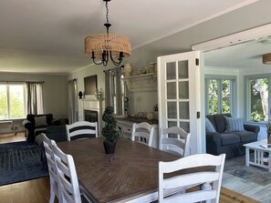 A dining area with a wooden table and white chairs, a chandelier, a fireplace, and a view through an open door to a living room. - Ponds Edge - waterfront -King bed -3 bedrooms 2 baths- kayaks - walk to beach (Barnstable)