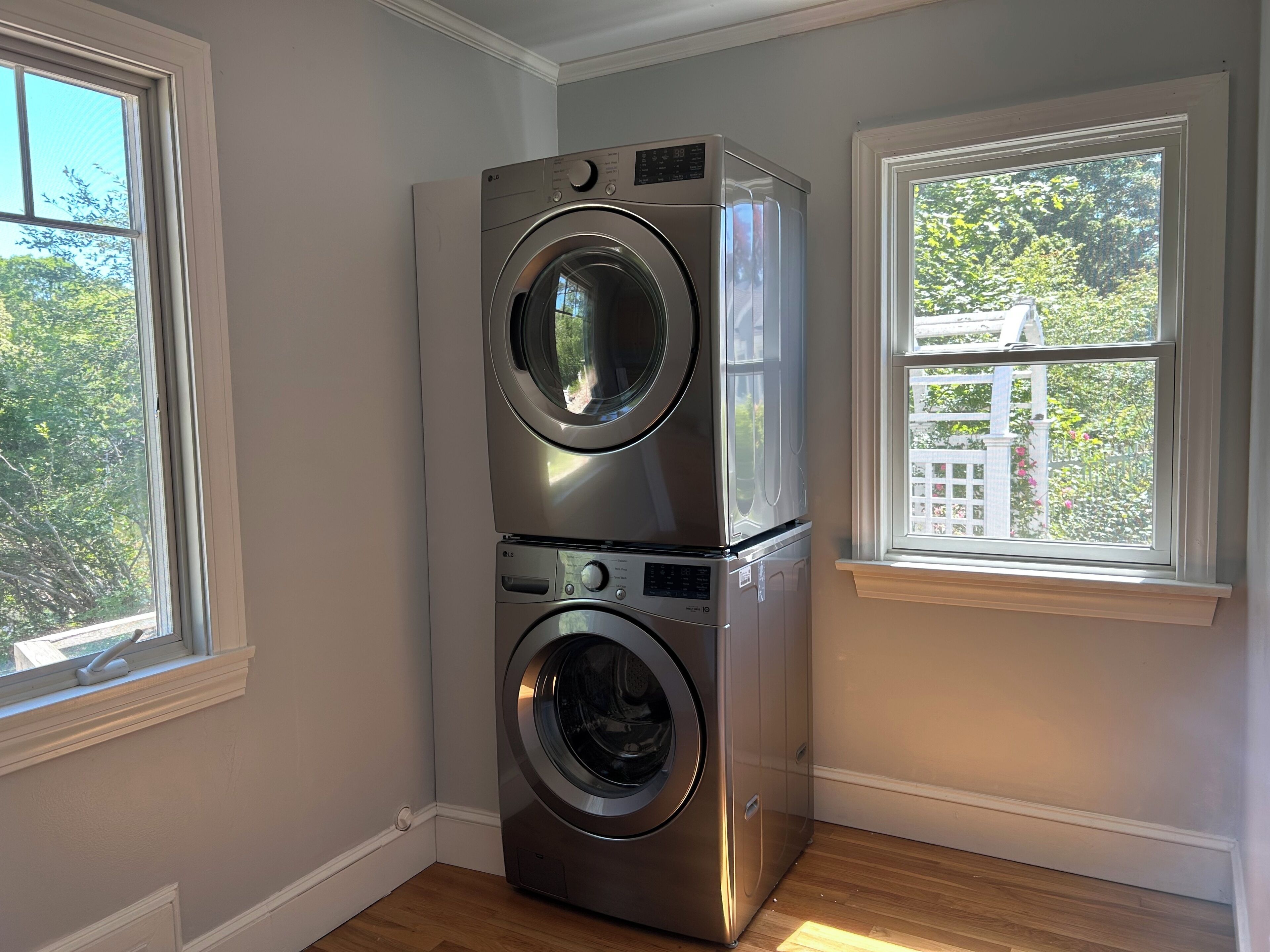 A stacked washer and dryer in a room with two windows, one of which has a view of greenery.