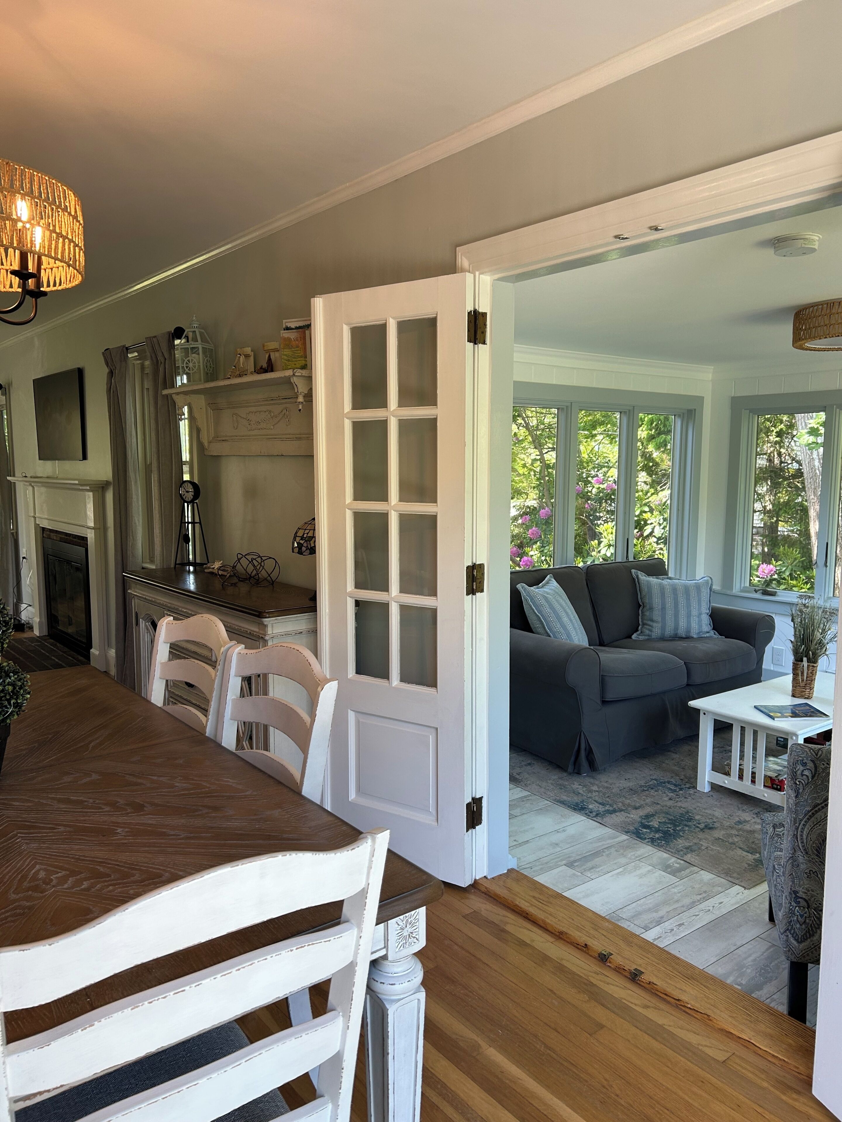 A dining area with a wooden table and chairs, a kitchen with a white door, a gray sofa, and a view of greenery outside.